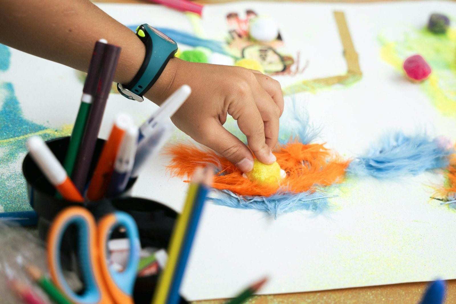 A child's hand creating a colorful art project with feathers and markers.